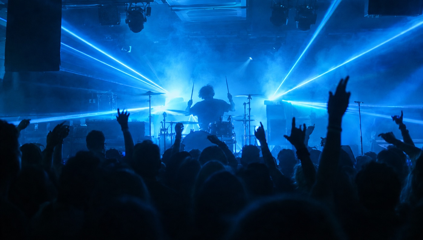 A club stage from the crowd, a drummer backlit by blue lasers in haze